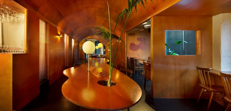 Vaulted timber ceiling over a curved dining table with palm fronds and globe pendant lights at dusk