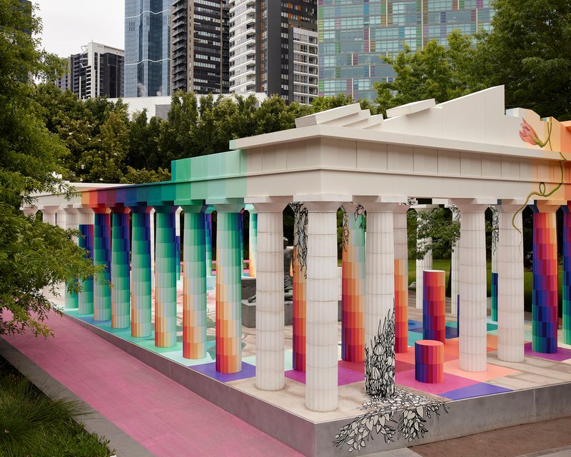 Corner view of the pavilion showing gradient columns, white tiled supports, and hanging foliage against urban towers