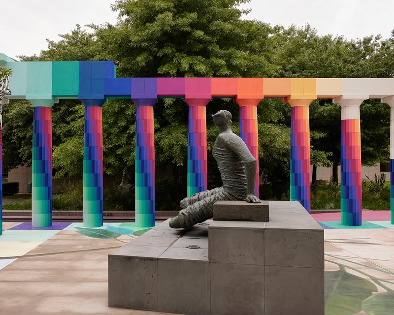Concrete sculpture of a seated figure on stepped plinth beneath the rainbow-gradient column capitals and beam