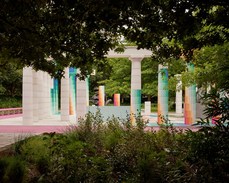 Open-air pavilion framed by mature tree canopy with pixelated columns and pink paving visible beyond