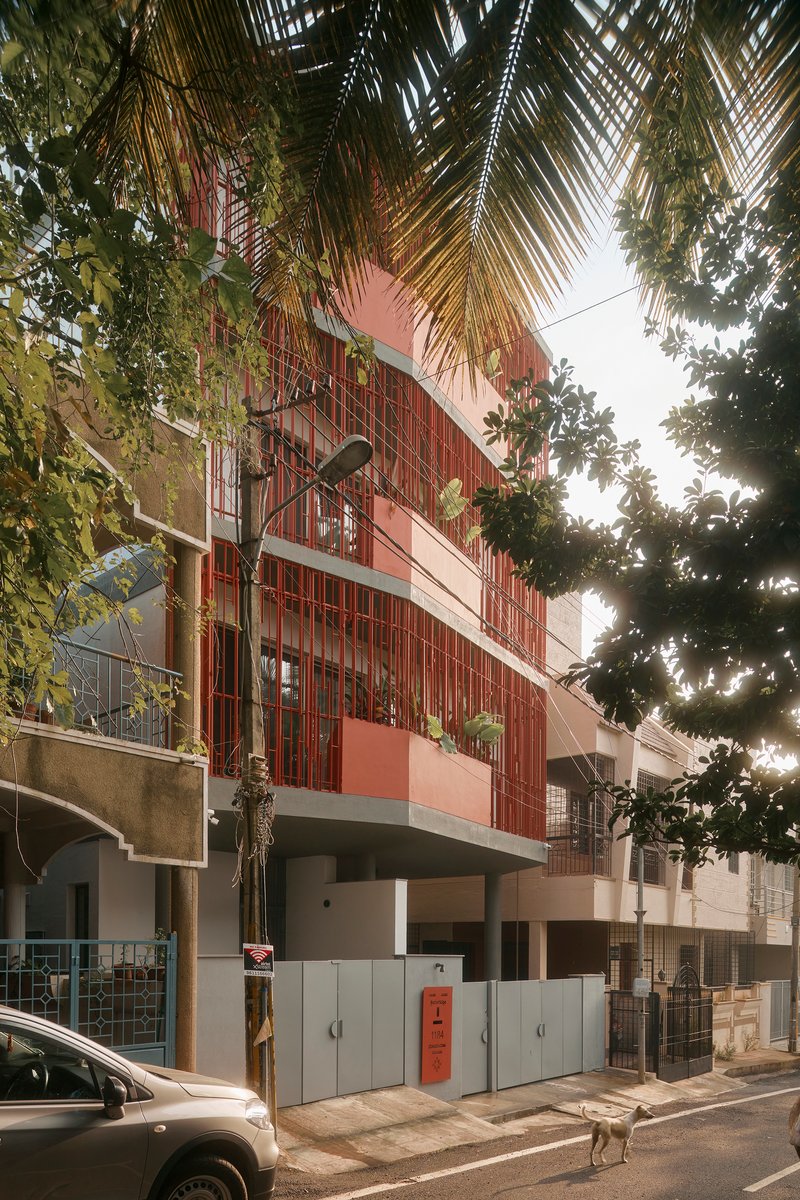 Street view of the red vertical batten facade rising above palm fronds and parked vehicles
