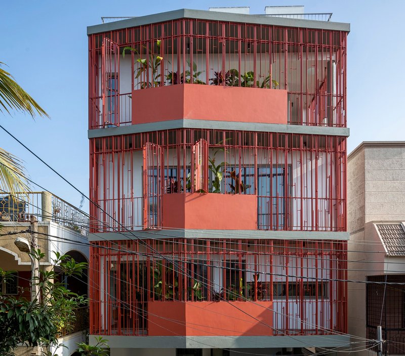 Street facade with red vertical metal brise-soleil wrapping three levels and planted terrace balconies