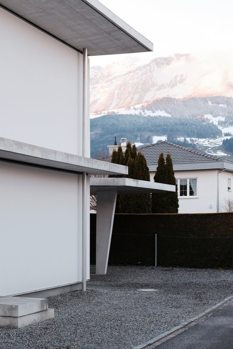 Concrete canopy supported by a slender column over a gravel drive with snowy peaks in the background