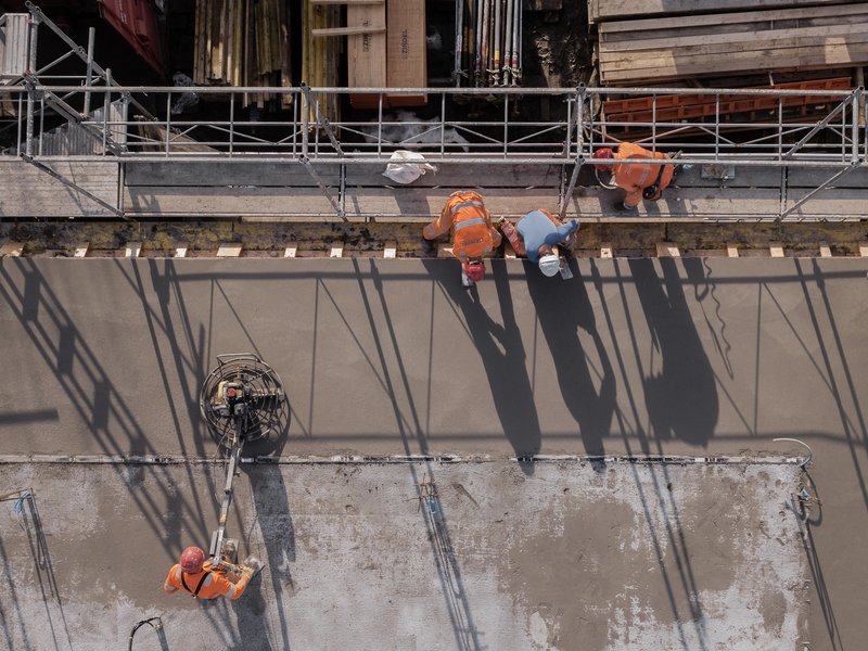 Construction workers on fresh concrete slab casting shadows through scaffolding above