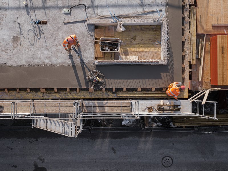 Drone view of construction workers positioning a timber-framed module beside fresh concrete slab