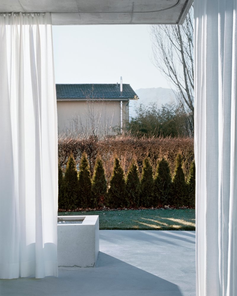 Covered terrace with sheer curtains and concrete bench overlooking a lawn edged by evergreen hedges