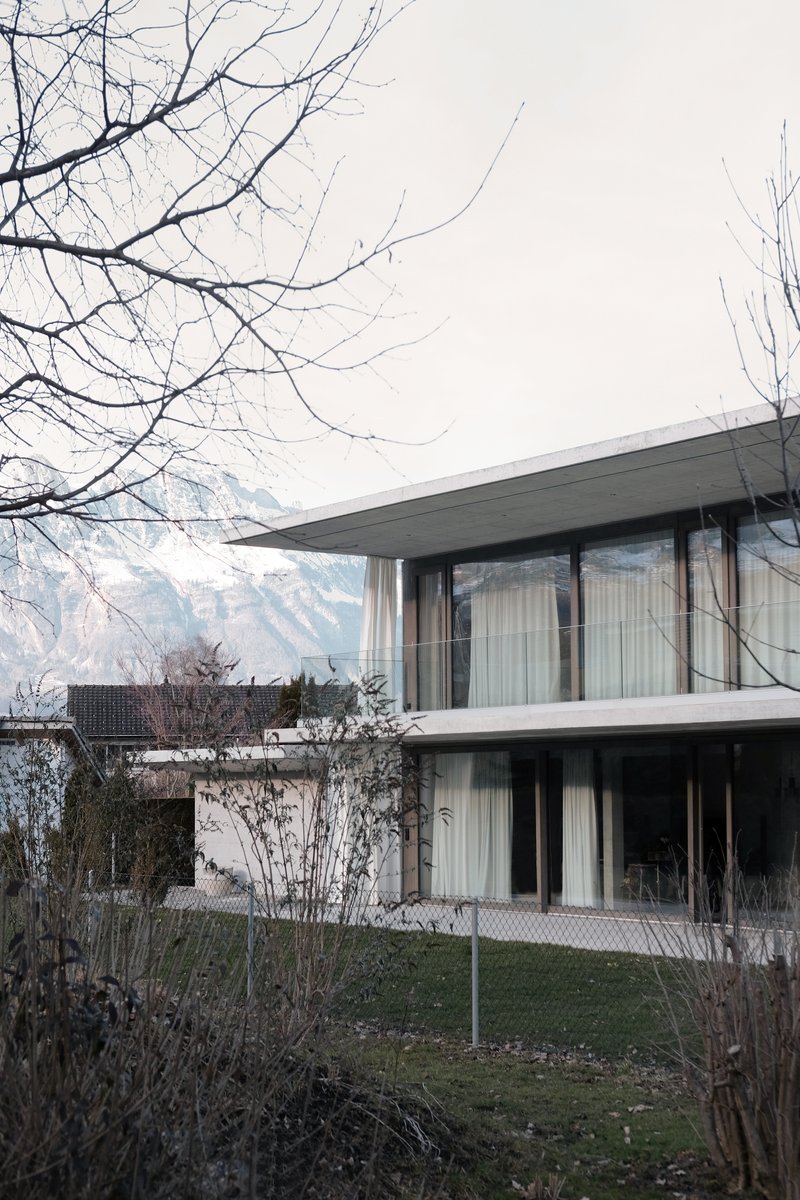 Two-story concrete volume with floor-to-ceiling glazing and sheer curtains framed by bare winter branches