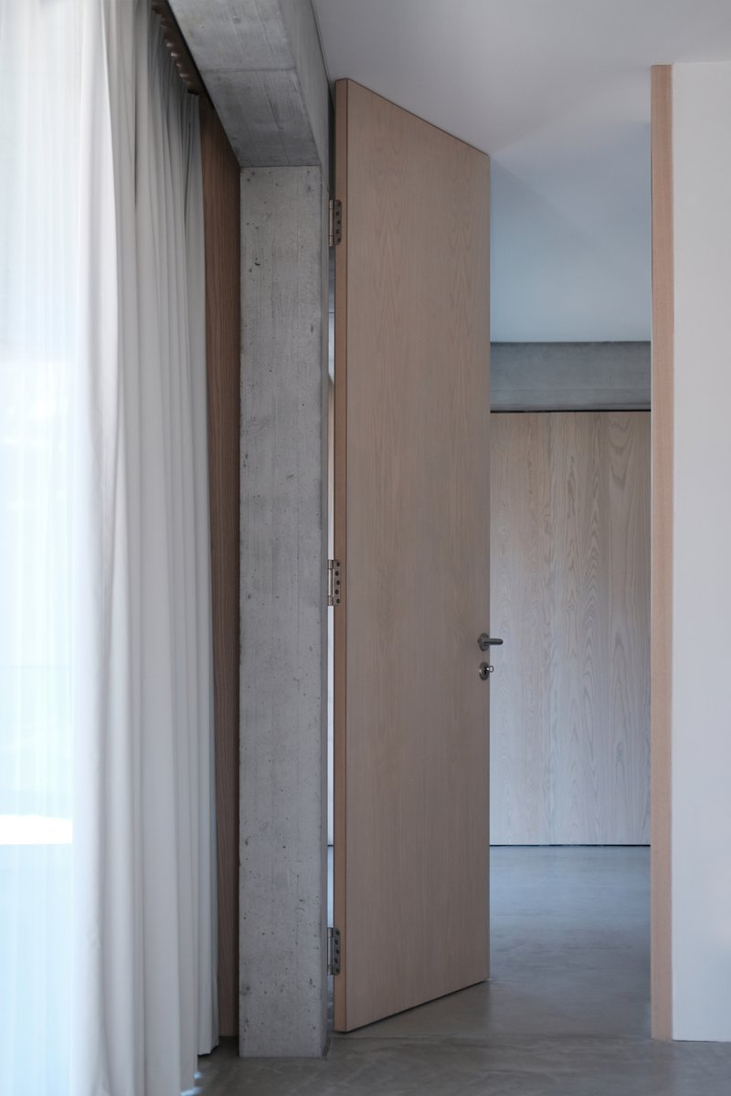 Interior hallway with light wood door ajar against concrete beam and white curtain at left