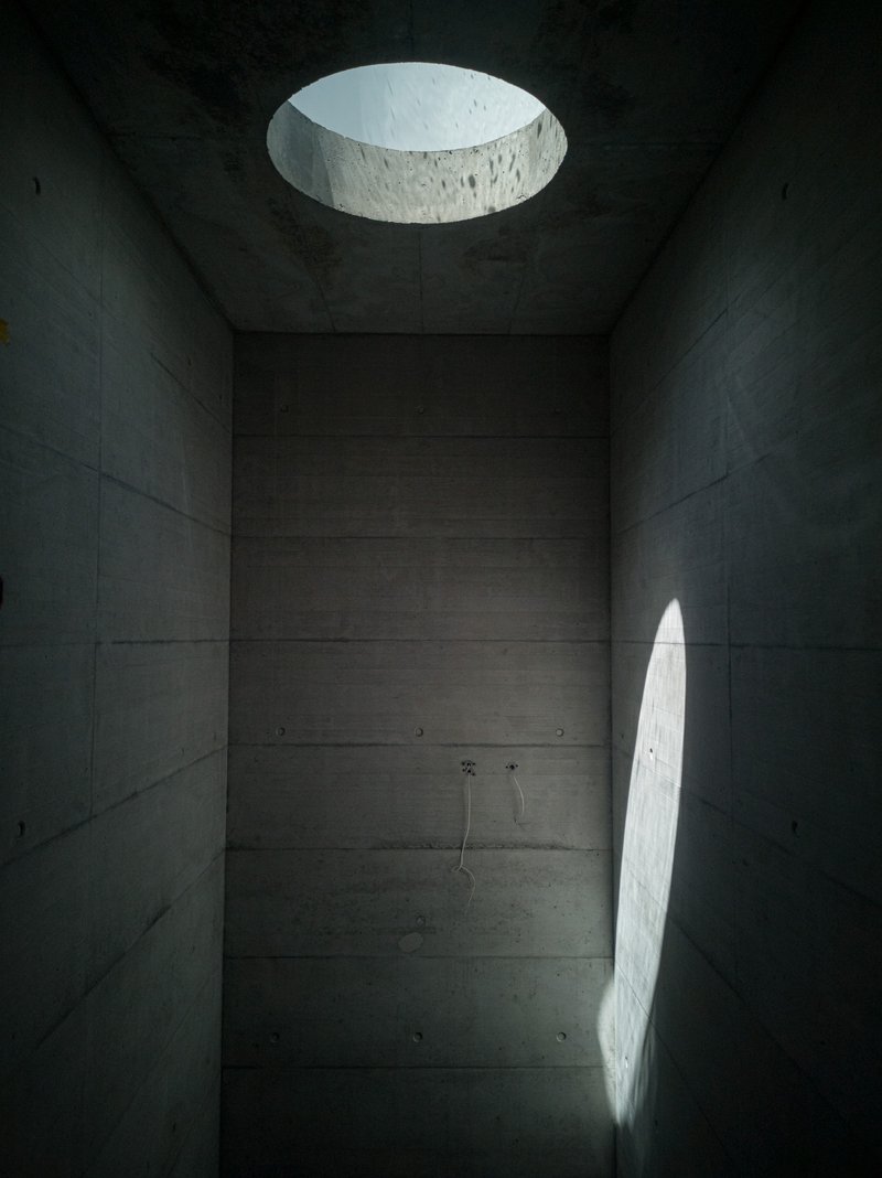Board-formed concrete bathing chamber lit by circular skylight and arched wall opening