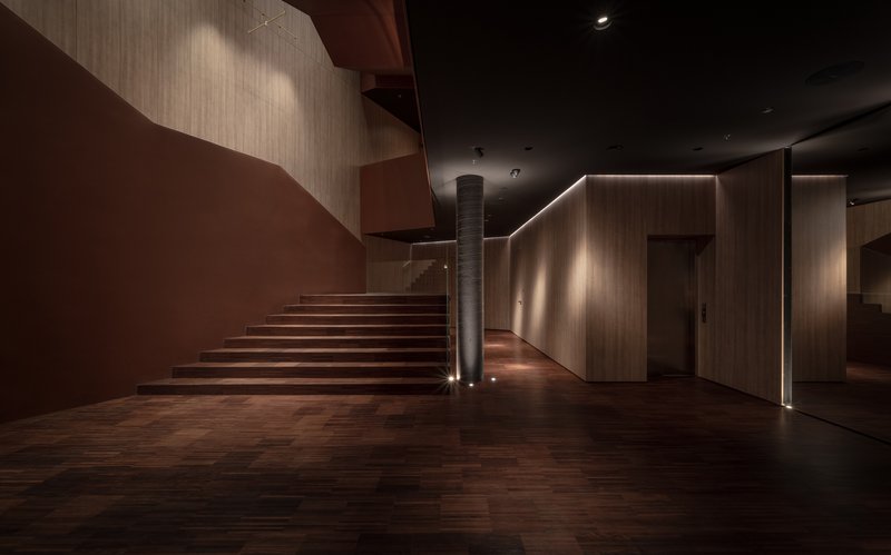 Cantilevered timber staircase rising from a wood-paneled lobby with recessed linear lighting and dark ceiling