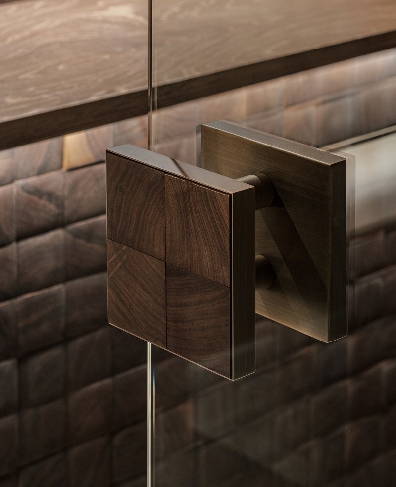 Close-up of a brushed metal door handle with walnut veneer plate against textured tile wall