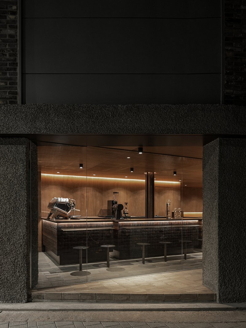 Night view through textured concrete portal into illuminated coffee bar with wood ceiling and tiled counter