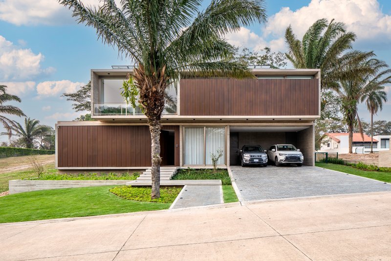 Street facade with vertical timber cladding, open carport, and palm trees under a cloudy sky