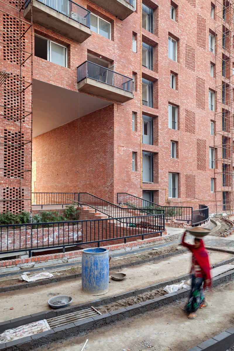 Red brick facade with perforated screens and recessed balconies, with a cyclist in motion on the construction site below