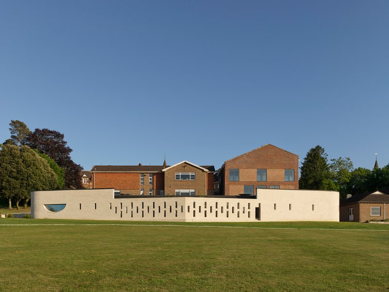 Pale concrete wall with vertical slit windows facing a green cricket pitch under clear blue sky