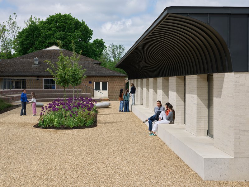 Courtyard with gravel surface and planted beds beside the ribbed metal canopy where visitors gather