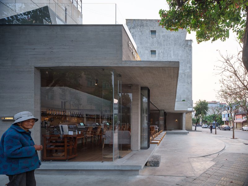 Corner glazed facade at street level with concrete canopy overhang and pedestrian passing under overcast evening light