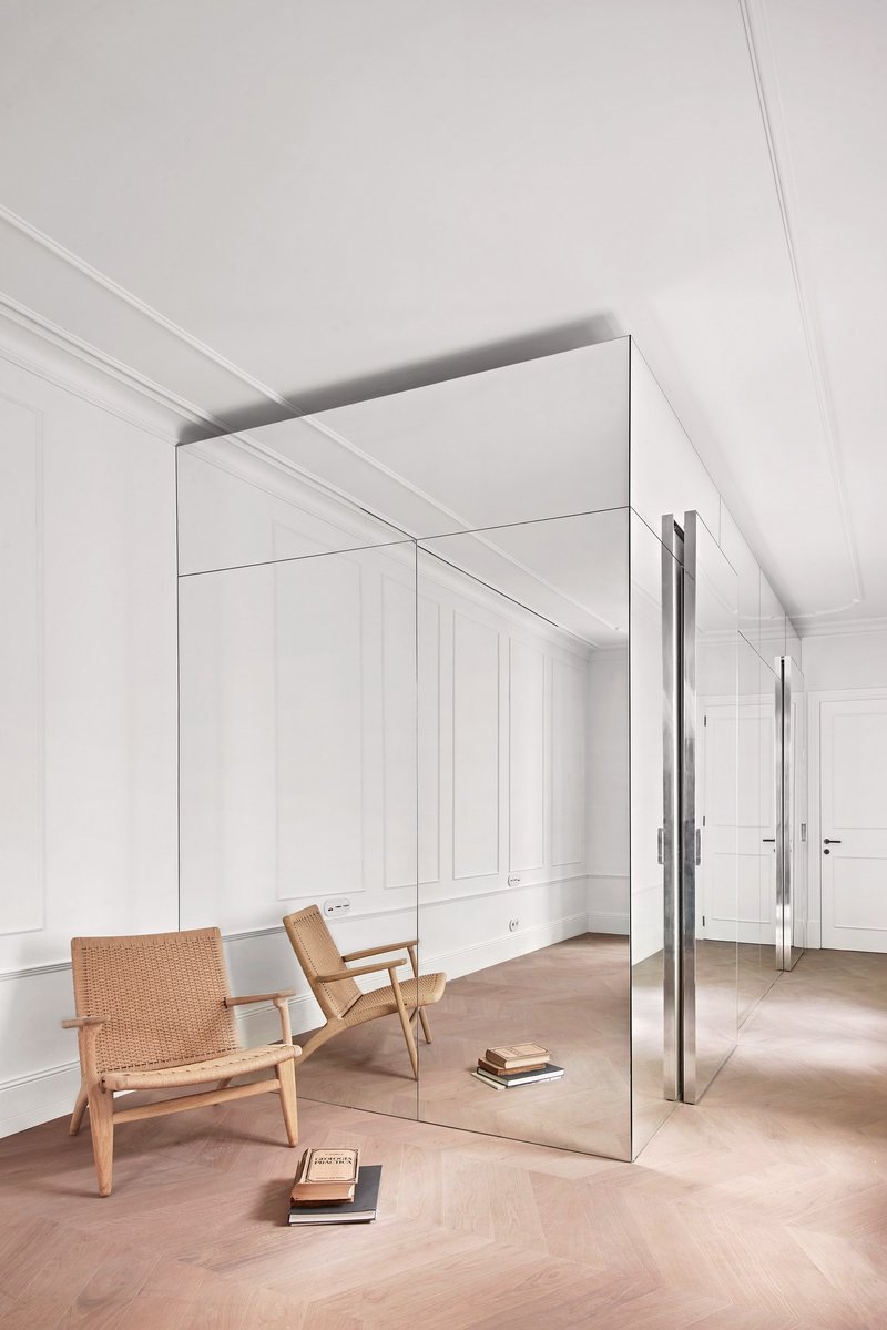 Freestanding mirrored volume with white paneled walls and herringbone oak flooring beside a woven chair