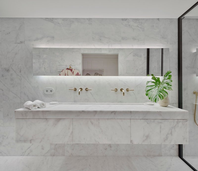 White marble vanity with integrated trough sink and brass wall-mounted faucets beneath a backlit mirror