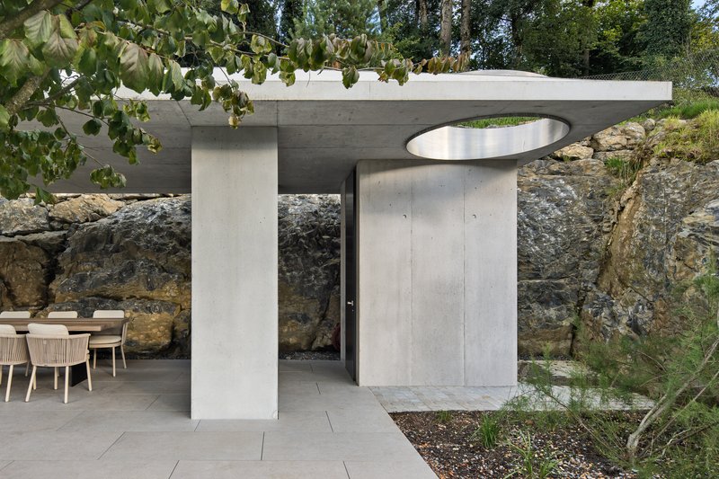 Concrete pavilion with flat roof and circular skylight opening set against exposed rock outcrop