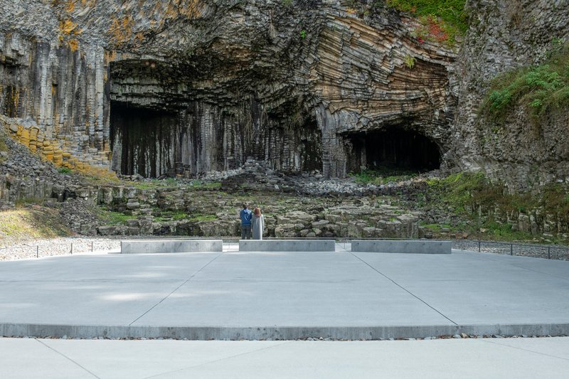 Concrete plaza with two visitors standing before the basalt cave openings and moss-covered walls