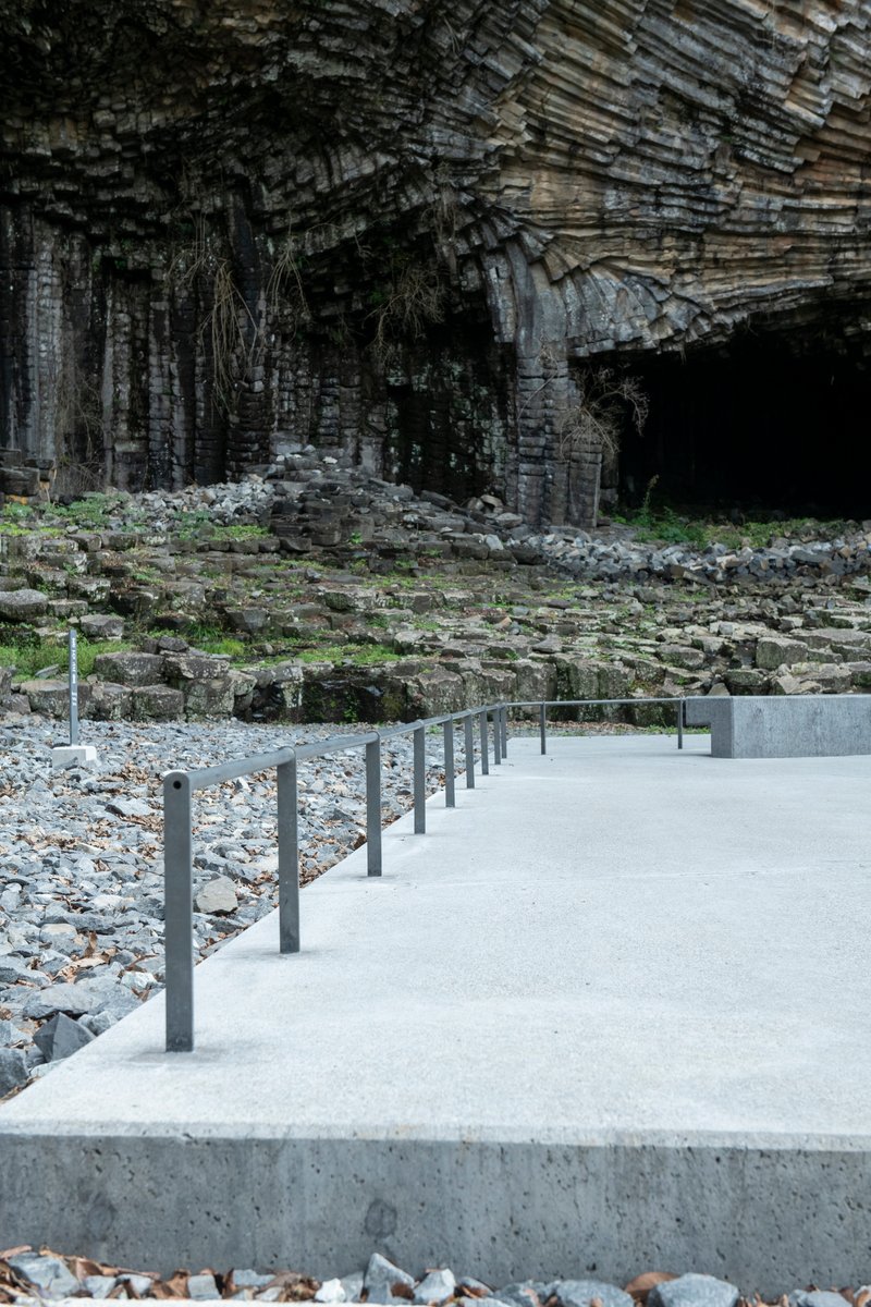 Concrete boardwalk with metal posts threading through a rocky shoreline below columnar basalt cliffs