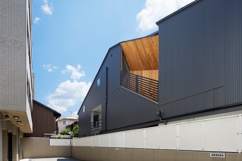 Dark corrugated metal facade with timber-lined balcony opening above a cream-colored perimeter wall under cloudy skies