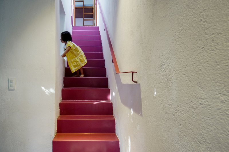 Child in a yellow dress ascending the maroon-painted staircase between white textured walls
