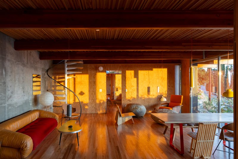 Living area with exposed timber beams and concrete wall beside spiral stair illuminated by warm evening light