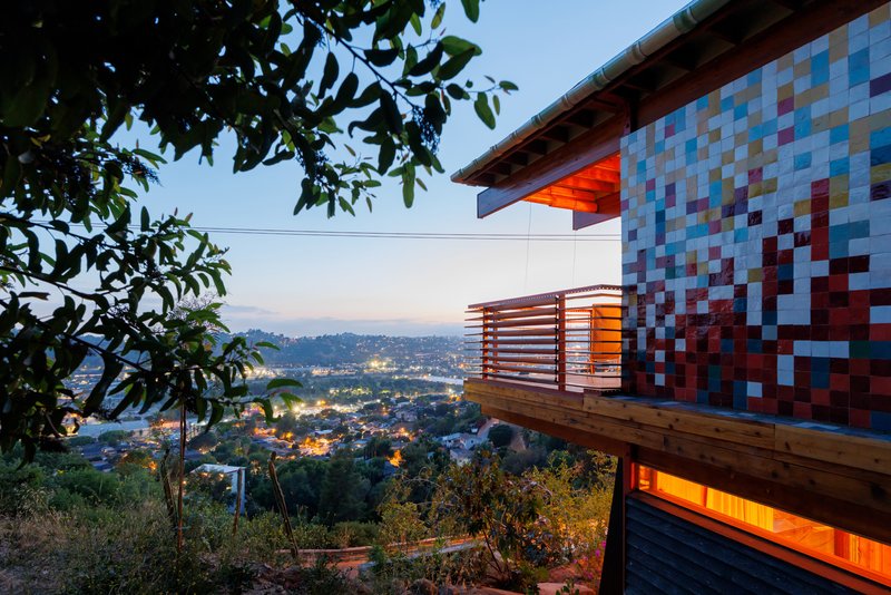 Cantilevered timber balcony with multicolored tile facade overlooking hillside neighborhood at twilight