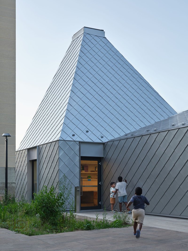 Diamond-patterned metal panel facade rising to a pyramidal volume as children run toward the entrance