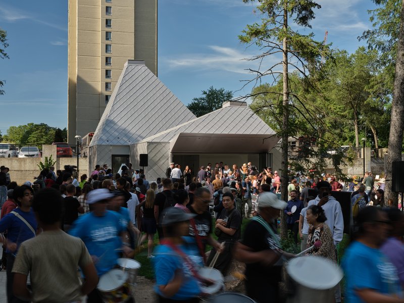 Gathered crowd surrounds the gray metal-clad pyramidal volumes in the afternoon light