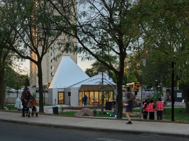 Pavilion with twin pitched roofs and open glass facade framed by pedestrians and park trees at dusk