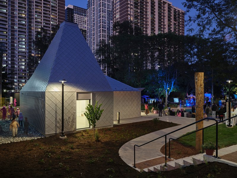 Illuminated pavilion with diamond-pattern metal facade and glazed openings at twilight among residential towers