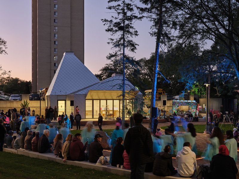 Crowd gathered on lawn steps facing the lit pavilion and park installations at sunset