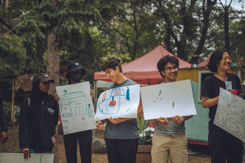 Group of participants holding architectural drawings outdoors beneath red canopy tents and mature trees