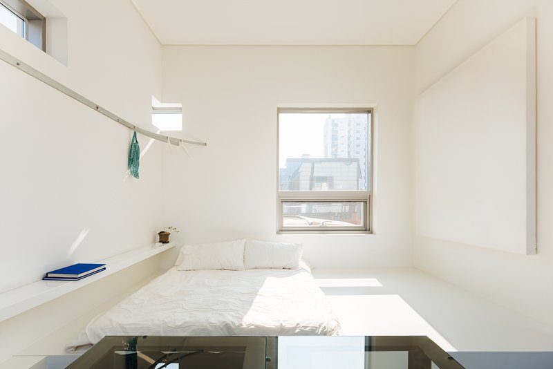 White bedroom with built-in bed platform and single window framing views of surrounding buildings