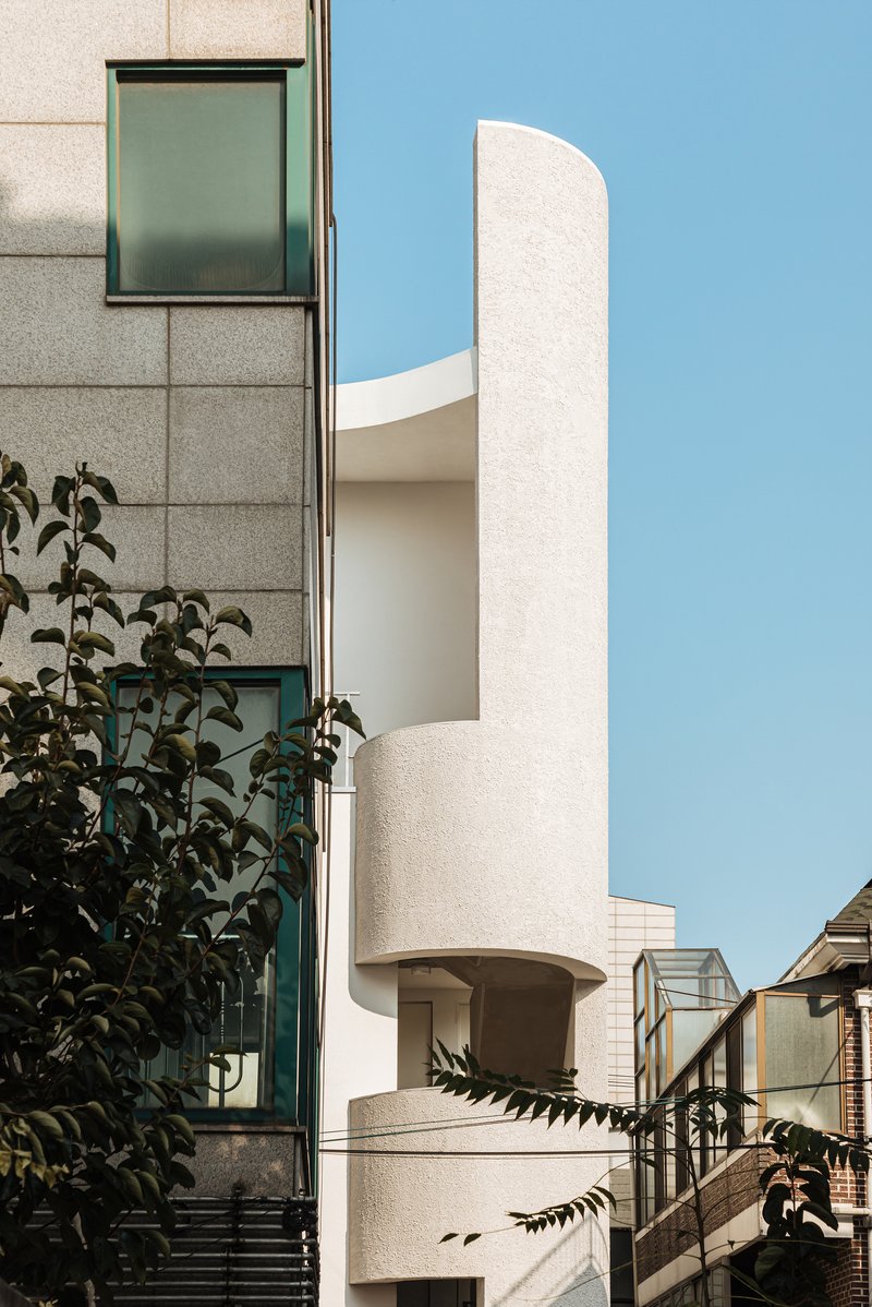 Textured cylindrical volume with inset balconies alongside neighboring building and foliage in foreground