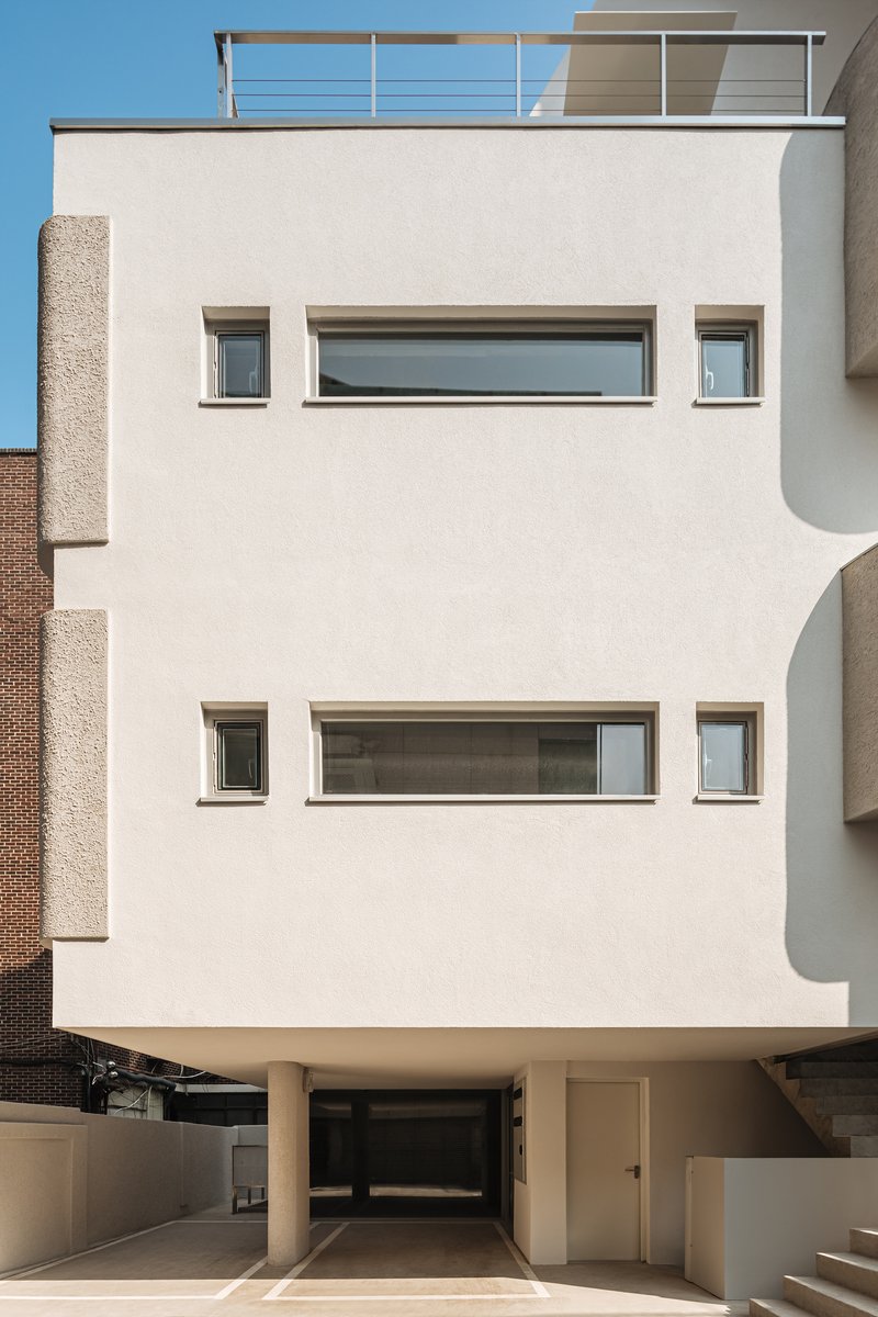 White stucco facade with horizontal strip windows and piloti supporting the raised volume under clear blue sky