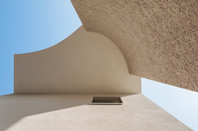 Upward view of curved stucco wall meeting rough textured surface with small skylight opening above