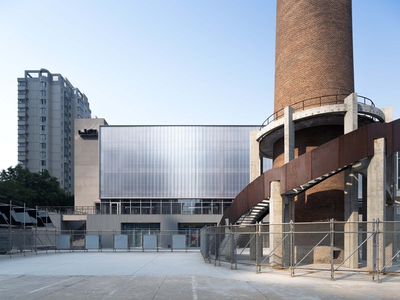 Translucent polycarbonate facade with a weathered brick chimney and elevated steel walkway at dusk