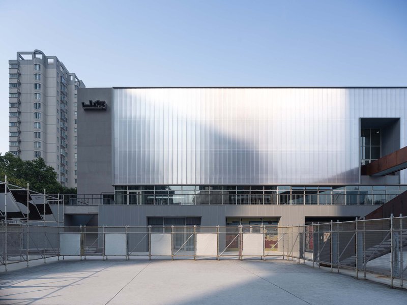 Vertical ribbed polycarbonate wall with horizontal window band above a concrete plaza with railings