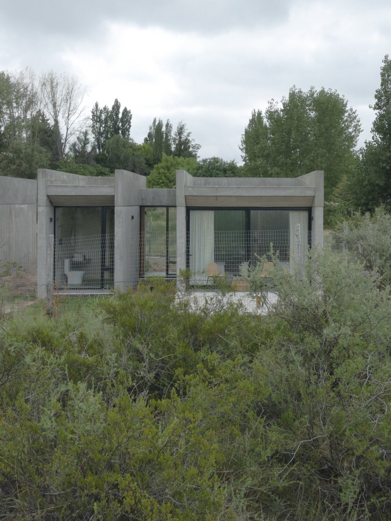 Front elevation showing the butterfly-roofed concrete structure among native scrubland under overcast skies