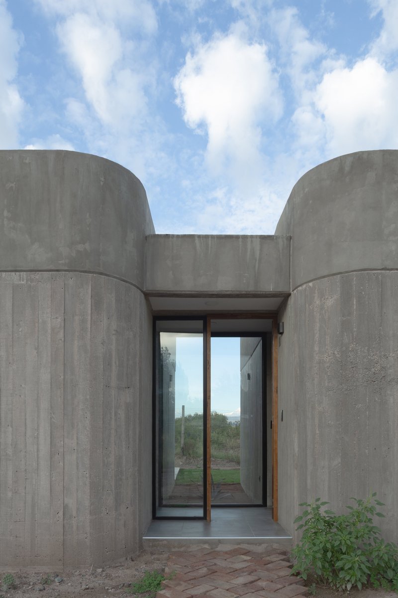 Glass entrance doors set between two cylindrical board-formed concrete volumes under a blue sky
