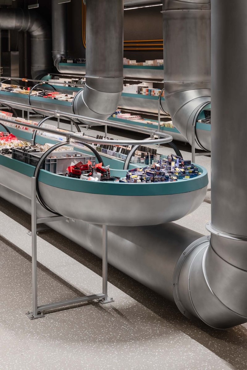 The image showcases an innovative retail space featuring a complex conveyor belt system for product display. The design incorporates industrial elements like me
