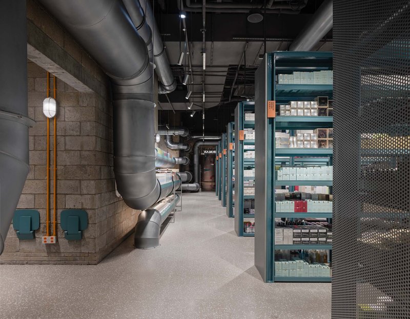 Corridor view with exposed ductwork running along a concrete block wall past teal shelving units
