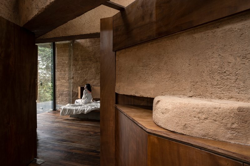 Curved wooden millwork meeting textured earthen walls with a figure seated on a bed beyond