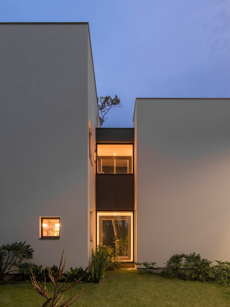Illuminated windows recessed into white volumes framing a small tree between planted beds at twilight
