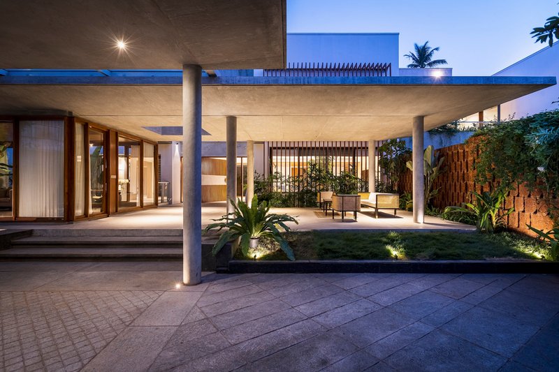 Covered outdoor terrace with concrete columns and slab roof at dusk, illuminated by uplights and tropical plantings