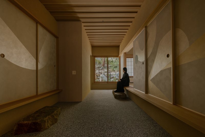 Narrow corridor with exposed timber ceiling beams and a seated figure facing a garden window